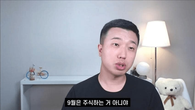 A man in a black shirt speaks directly to the camera in a room featuring a floor lamp, decorative bicycle, and teddy bear, with Korean subtitles reading "9월은 주식하는 게 아냐" ("September is not for trading stocks"), reinforcing the post's warning against stock investing in September.