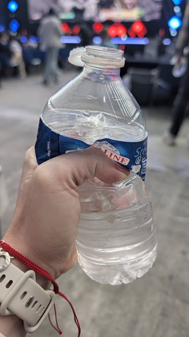 A hand with a white Apple Watch band holding a partially crushed plastic water bottle at what appears to be a stadium or arena event.