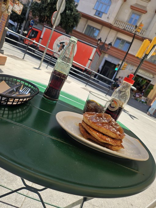A casual outdoor café scene with a plate of toasted bread, a glass of Coca-Cola, and a glass bottle on a green table, with a street and buildings visible in the background.