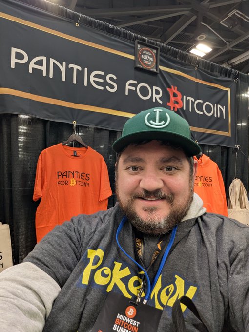 A man wearing a green cap and a Pokémon t-shirt taking a selfie in front of a booth with a banner that reads 'PANTIES FOR BITCOIN'.
