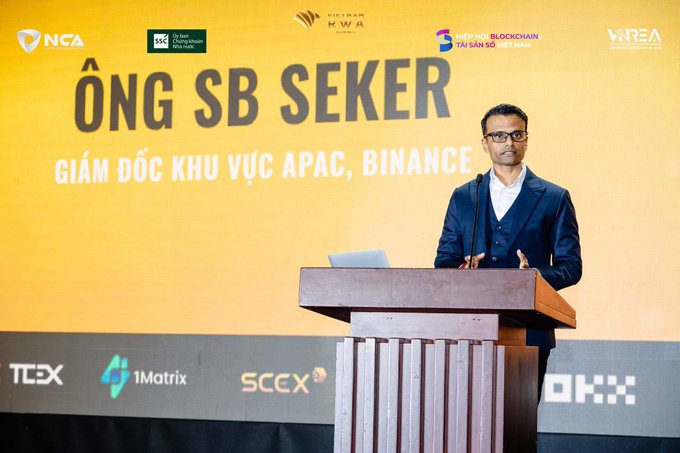 A man in a suit and glasses speaking at a podium during a conference, with a yellow backdrop displaying 'ÔNG SB SEKER', 'GIÁM ĐỐC KHU VỰC APAC, BINANCE', and various sponsor logos.