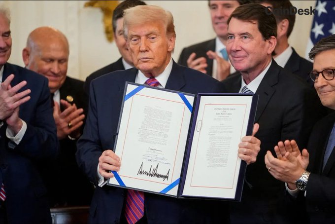 President Donald Trump holds a signed document, as lawmakers behind him applaud, likely representing the passage of legislation. This image suggests a legislative victory, connecting to the news about the CLARITY Act's potential floor vote.
