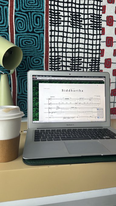 A MacBook Air sits open on a desk displaying sheet music titled 'Siddhartha', accompanied by a takeaway coffee cup and a desk lamp in front of a patterned tapestry.