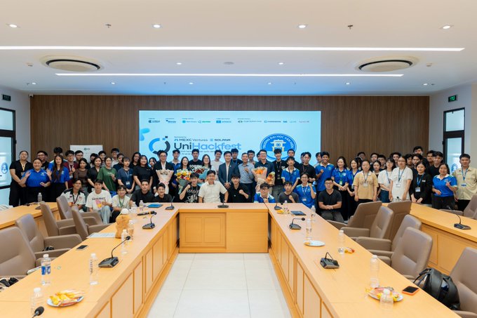 A large group of students and event organizers gather for a group photo in a modern university conference room, posing in front of a screen displaying the "Uni-Hackfest" banner held at Van Hien University (VHU) in partnership with MEXC Ventures. The "Technology Capital Flow & Investment Thinking" seminar brought together students and speakers for an engaging session on finance and investment.