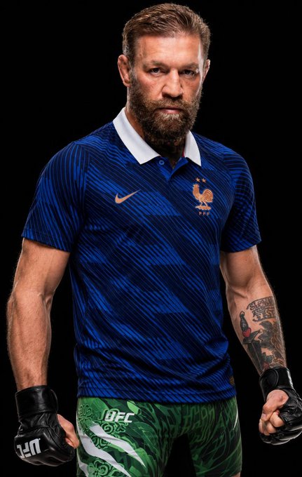 A man with a beard wearing a blue France national team Nike jersey and green UFC fight shorts, with black UFC gloves on his hands.