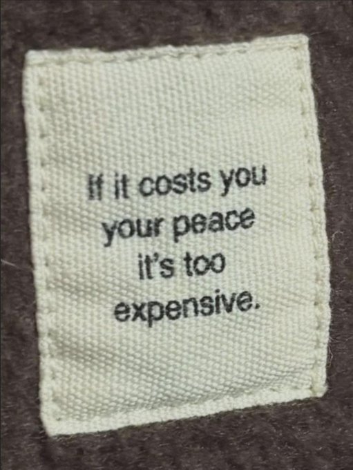 A small cloth label sewn onto brown fabric displays the printed quote: "If it costs you your peace it's too expensive." The message reinforces the post's idea of determining your cost—suggesting that any price paid at the expense of inner peace is not worth it.