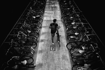 A black and white photograph captures a lone cyclist from above, riding down a path flanked by rows of parked bicycles, with the number "7" painted on the pavement. The image, titled "Street Vibes," evokes the solitude of urban commuting and is highlighted as part of the #TezosTuesday NFT drop on @objktcom.
