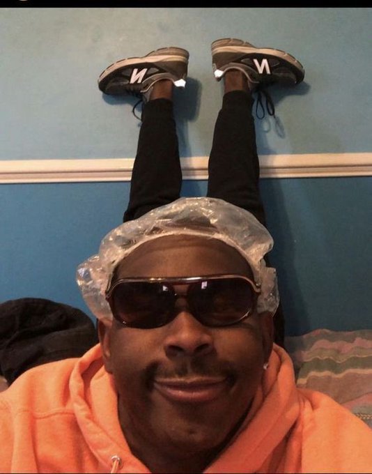 A man wearing sunglasses, a shower cap, and an orange hoodie lies upside down with his legs propped against a blue wall, sporting "N" and "V" lettered sneakers. The playful upside-down pose fits the casual "gm gang" good morning greeting vibe.