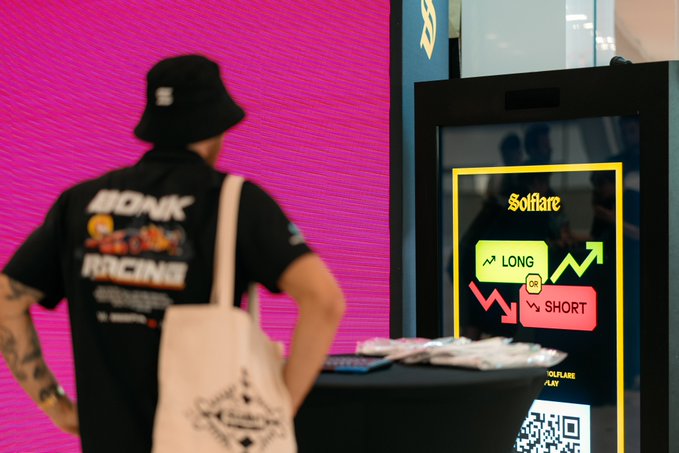 A person wearing a Bonk Racing shirt and a tote bag stands before a Solflare kiosk at the Solana Summit, where a digital display prompts attendees to choose between "LONG" and "SHORT" options alongside a QR code—visually capturing the playful "long or short?" dilemma highlighted in the post.