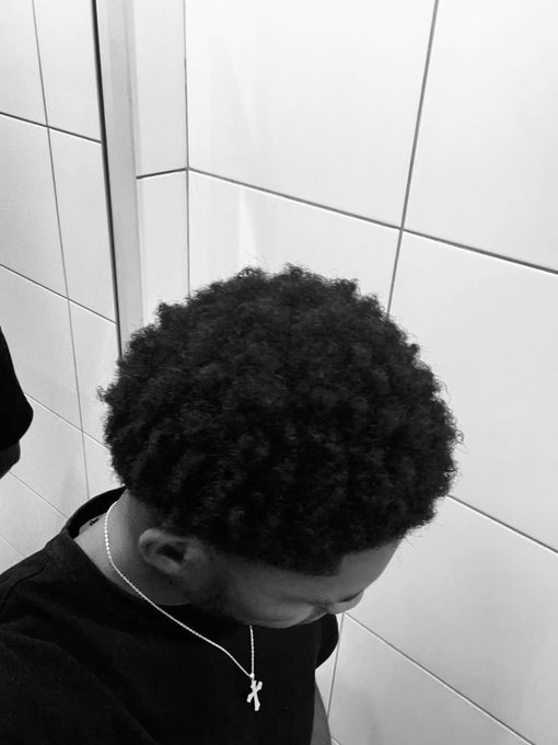 A black and white selfie taken from above showing a young man with a curly afro haircut, looking toward the ground while wearing a silver cross necklace and black shirt against a white tiled wall.