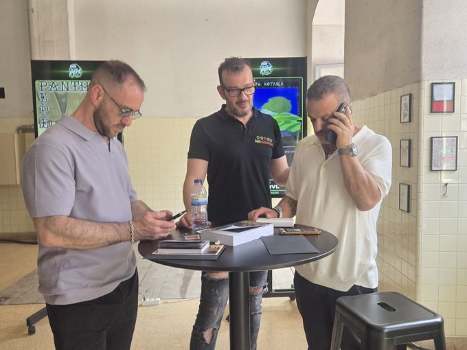 The image shows three men standing around a table with papers and books on it. One man is holding a cell phone, possibly taking notes or discussing something related to their work at the PEPEVAULT booth. The other two men are engaged in conversation as well, likely sharing ideas or discussing their projects.