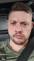 A close-up selfie of a man with short brown hair and a beard wearing an olive green t-shirt and seatbelt, sitting inside a vehicle.