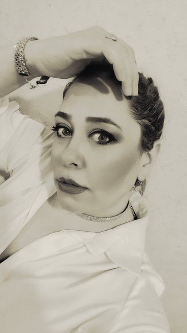 A sepia-toned portrait of a woman with dramatic eye makeup resting her hand on her head, wearing a white shirt and jewelry.