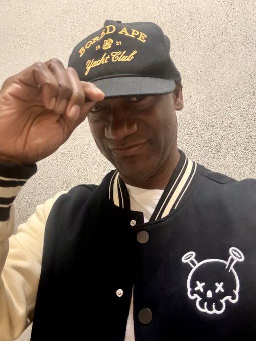 A man wearing a black "Bored Ape Yacht Club" cap and a black and cream varsity jacket smiles while tipping his hat.