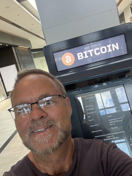 A selfie of a smiling man with glasses standing in front of a Bitcoin ATM displaying the orange 'BITCOIN' logo.