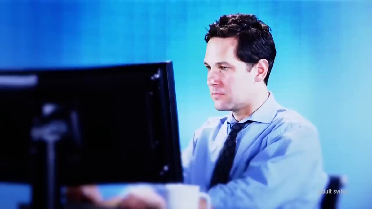 The image shows a man sitting at his desk, wearing a blue shirt and tie, with a computer monitor in front of him. He has a cup of coffee next to him as he works or studies. The background features a blue wall that is slightly blurred, drawing attention to the main subject - the man working on his computer.
