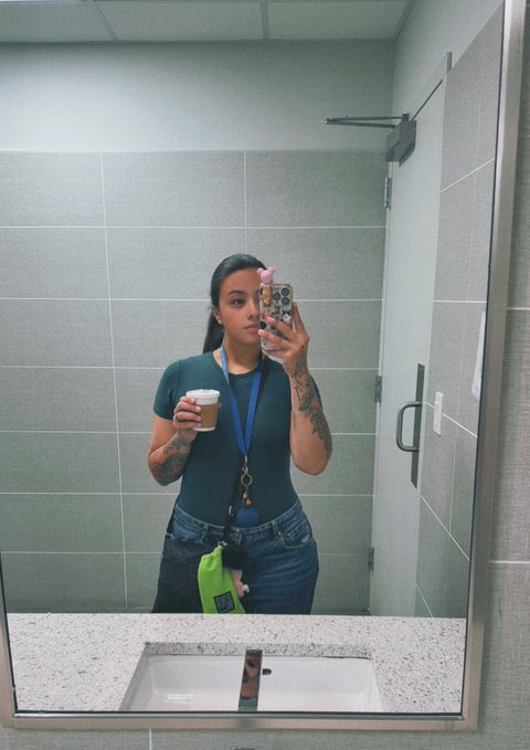 A woman with tattoos takes a mirror selfie in a bathroom while holding a coffee cup, wearing a lanyard with keys.