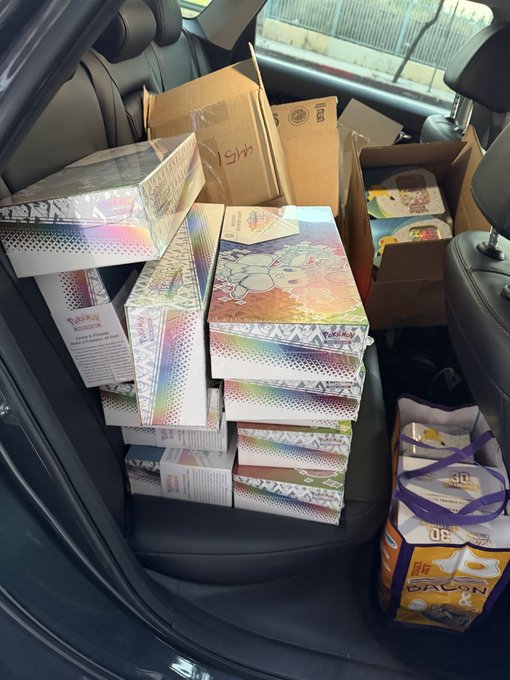 The back seat of a car is overflowing with stacks of Pokémon Trading Card Game products, including tins, boxes, and a bag of Ecruteak ("90..") booster packs, likely headed to or from a card shop, trade, or tournament. It perfectly captures the chaotic "average day" of a dedicated Pokémon TCG collector or reseller.