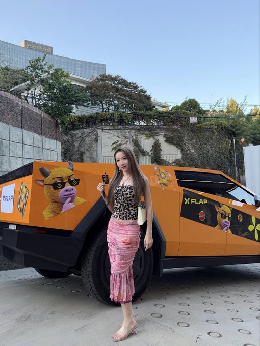 A young woman in a leopard print top and pink skirt posing in front of a Tesla Cybertruck wrapped in a bright orange "X FLAP" advertisement featuring a cartoon cow character.
