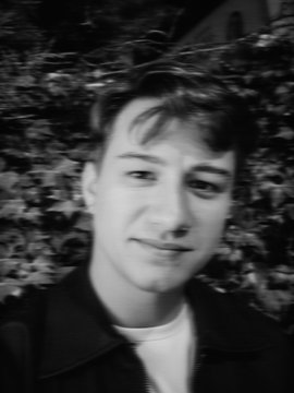 A black and white portrait of a young man standing outdoors with foliage in the background.