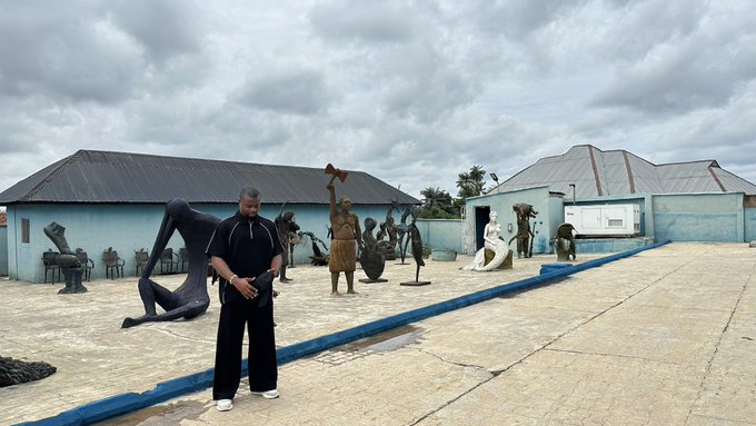 A man in dark clothing standing in an outdoor sculpture garden displaying various artistic statues, including figures with animal and human forms, under a cloudy sky.
