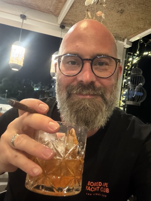 A bald, bearded man wearing glasses holds a glass of amber liquid with ice and a cigar, wearing a black shirt with 'BORED APE YACHT CLUB' branding.