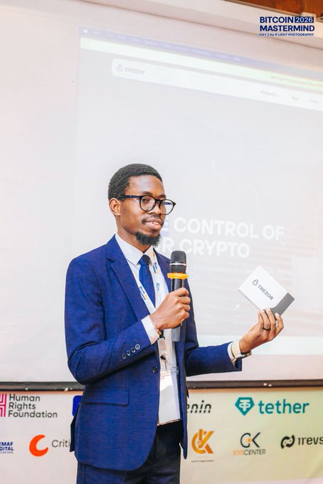 A man in a blue checkered suit and glasses speaks into a microphone while holding a Trezor hardware wallet at the Bitcoin 2026 Mastermind event, with a presentation screen reading 'CONTROL OF YOUR CRYPTO' behind him and sponsor logos including Tether.