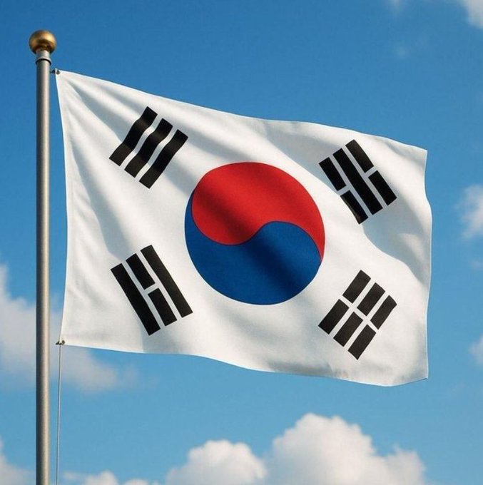 The South Korean flag, featuring the red and blue taegeuk symbol and four black trigrams on a white background, waves on a flagpole against a blue sky with clouds.