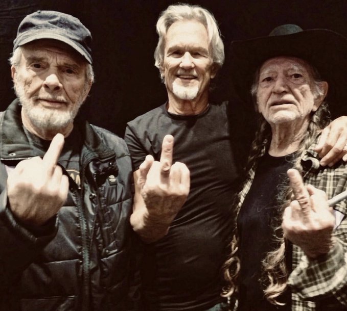Three older men pose together for a photo, each raising their middle finger toward the camera in a rebellious gesture, with the man in the center identified as Kris Kristofferson.
