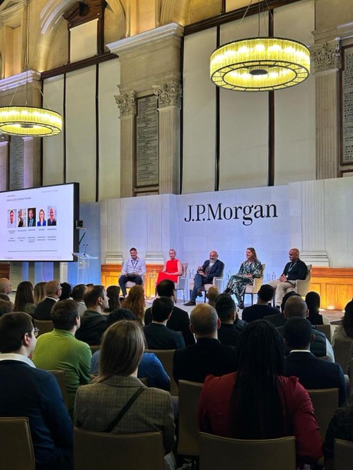 A J.P. Morgan panel event taking place in a grand hall with chandeliers. Four panelists are seated on stage in front of a J.P. Morgan backdrop, with an audience watching. A screen on the left displays 'JPMorgan', 'Driving Inclusive Finance', 'Panel Discussion', and headshots of speakers.