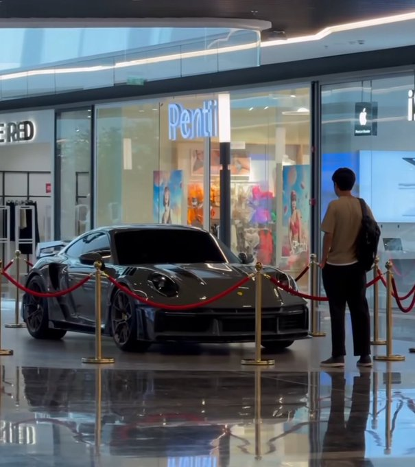 A sleek black sports car is on display behind red velvet ropes in a modern mall, with a young man in a tan shirt and backpack pausing to admire it, while signs for "Pentil" and "RED" are visible in the background. The image symbolizes aspirational achievement and the motivation to work toward owning such a luxury vehicle, reinforcing the message "Your time is coming. Believe."