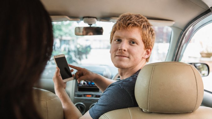 A young man with red hair sits in the driver's seat of a car, holding up a smartphone with a dark image on the screen while looking back at a woman in the passenger seat.