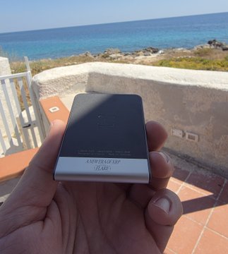 A hand holds an 'iPhone 13 Pro Jet Black' box upside down in front of a scenic ocean view from a balcony.