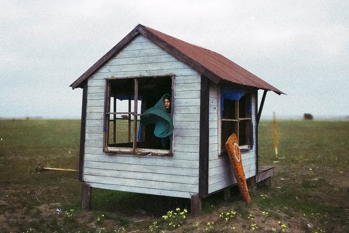 A small white wooden shack with a red roof sits alone in a flat grassy field under an overcast sky, with a figure in a green hooded garment looking out the window.