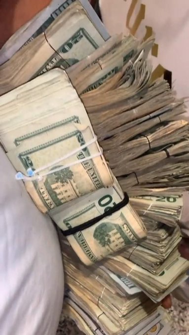 A person's hand holds a thick bundle of US currency, primarily $20 bills, bound together with rubber bands.