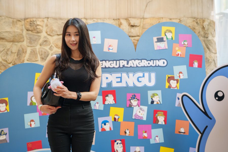 A smiling woman in a black outfit poses at the "Pengu Playground" event backdrop, holding a small pink plush penguin, in promotion of Pudgy Penguins.