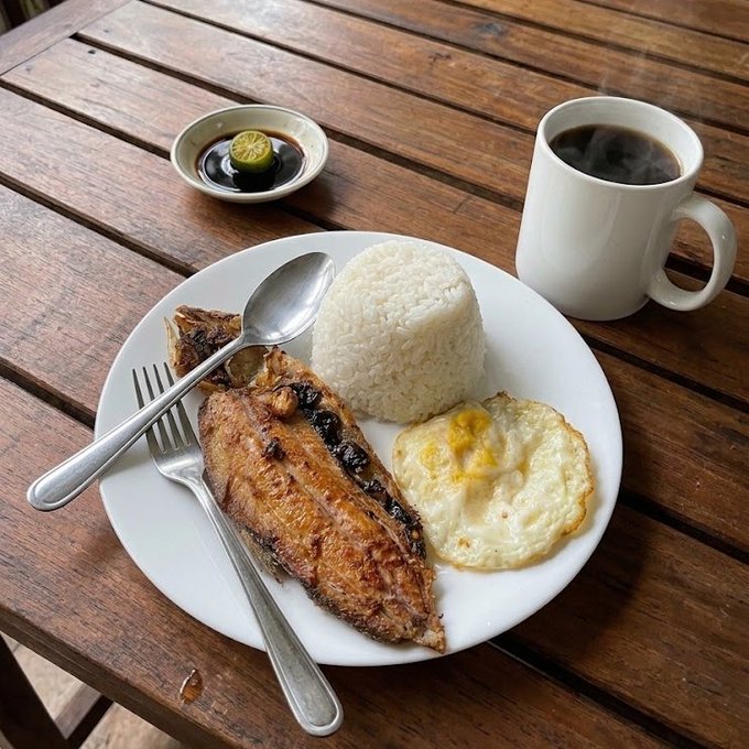 A Filipino-style meal served on a wooden table, featuring grilled fish, white rice, a fried egg, a small dish of soy sauce with calamansi, and a mug of steaming coffee.
