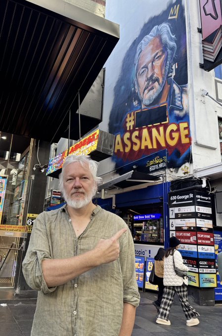 A white-bearded man points to a large mural of himself with the text '#ASSANGE' on a city building.