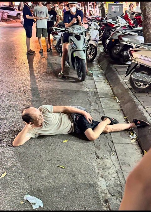 A man lies relaxed on a sidewalk, seemingly oblivious to the bustling street scene around him where a traffic officer checks his phone and onlookers gather near a row of parked motorbikes, perfectly capturing the "laying down on the job" energy the post humorously wishes upon its followers.