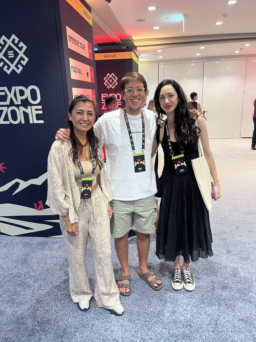 Three people posing together at a convention in front of an 'EXPO ZONE' sign, with visible stage names including 'RIVERDALE STAGE' and 'FOOTBALL', and signage indicating 'EXPO 2024'.