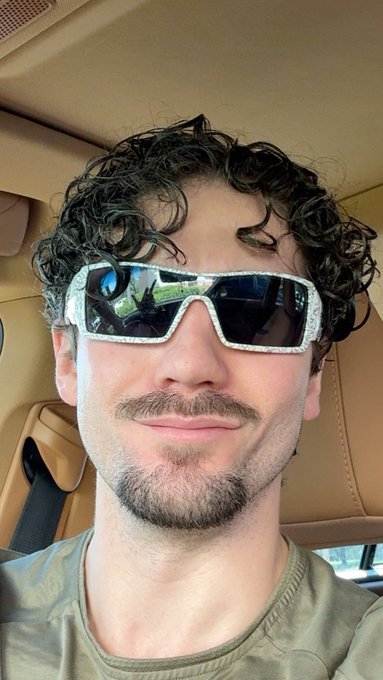 A man with curly hair and a subtle smirk sits in a car wearing oversized, sparkly rhinestone sunglasses, projecting a confident and playfully luxurious vibe that matches his caption about getting great sleep and being on the verge of making "generational wealth."