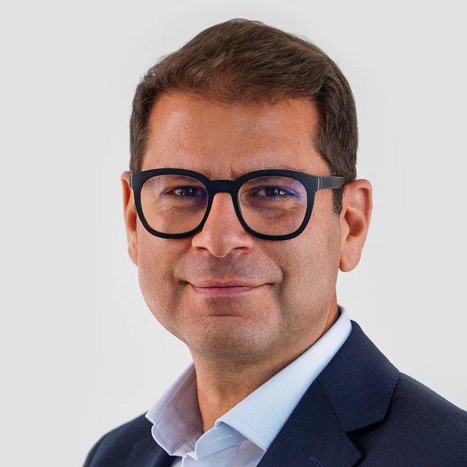 A professional headshot of a man with short brown hair, wearing black-rimmed glasses, a dark suit jacket, and a light blue shirt against a white background.