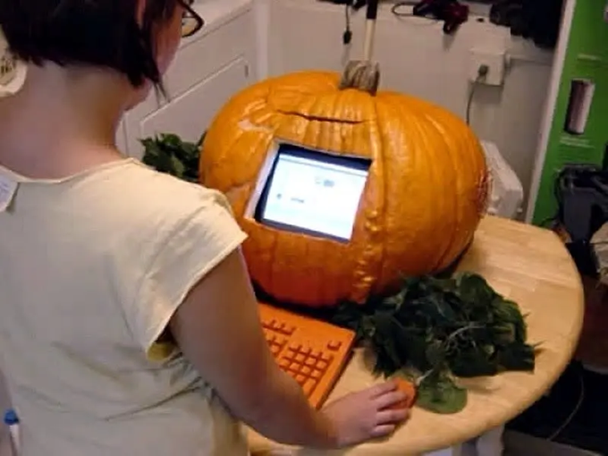 A child uses an orange keyboard and mouse to interact with a laptop embedded in a large pumpkin, with a small pumpkin serving as a mouse.