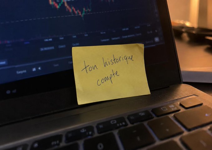 A yellow sticky note with the French handwritten text "ton historique compte" is stuck to the screen of a laptop displaying financial candlestick charts.