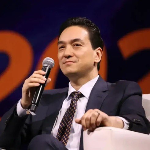A man in a dark suit, white shirt, and patterned tie holds a microphone while seated at what appears to be a panel or conference event.