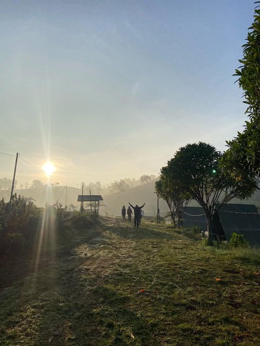 A silhouette of a person with raised arms stands on a misty dirt trail at sunrise, with a few other figures and a small shelter visible in the foggy distance.