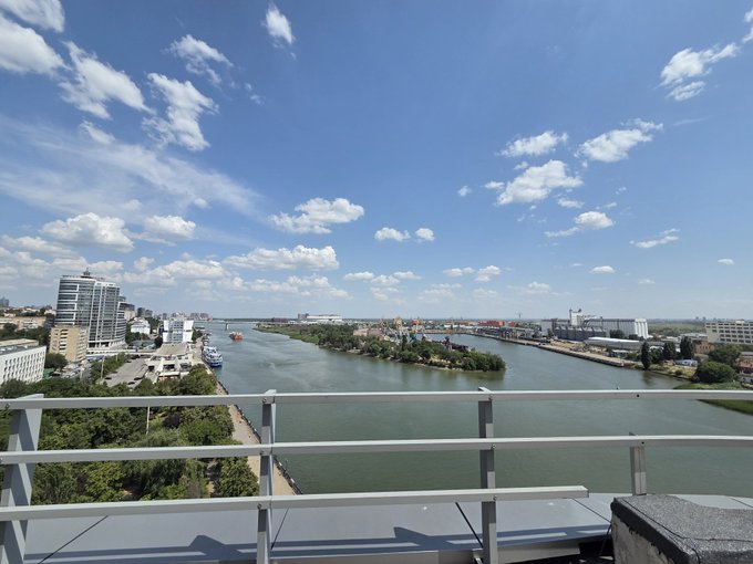 A daytime cityscape photograph taken from a balcony, overlooking a wide river, an island, and urban buildings under a blue sky with scattered clouds.