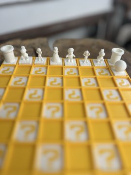 The image features a yellow chess set with white squares, including a king piece positioned at the center. The chess pieces are posed for a photo, showcasing their unique designs. In the background, there's a blurred image of a person sitting on a chair, which adds context to the scene and suggests that this could be an advertisement or promotional material related to the chess set.