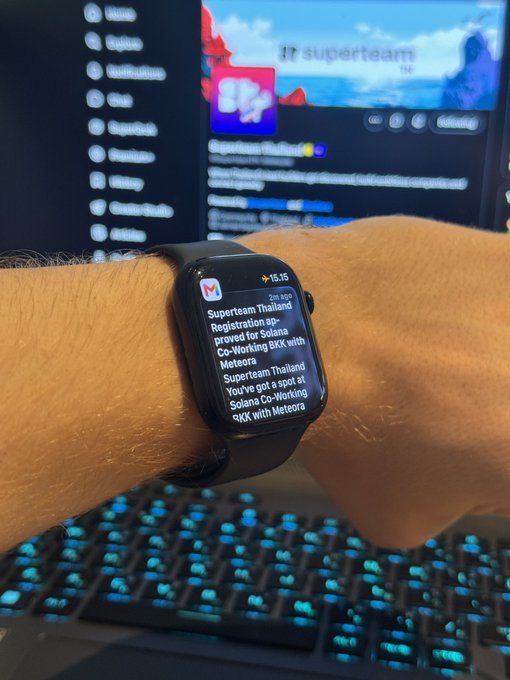 A wrist wearing an Apple Watch displaying a Gmail notification about 'Superteam Thailand Registration approved for Solana Co-Working BKK with Meteora', with a glowing keyboard visible below.