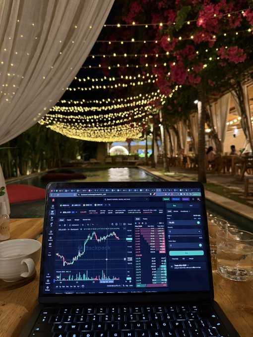 A laptop displaying a cryptocurrency trading chart with 'Backpack' branding sits open on a wooden table at an outdoor evening venue, surrounded by string lights, bougainvillea flowers, white curtains, and a swimming pool in the background.